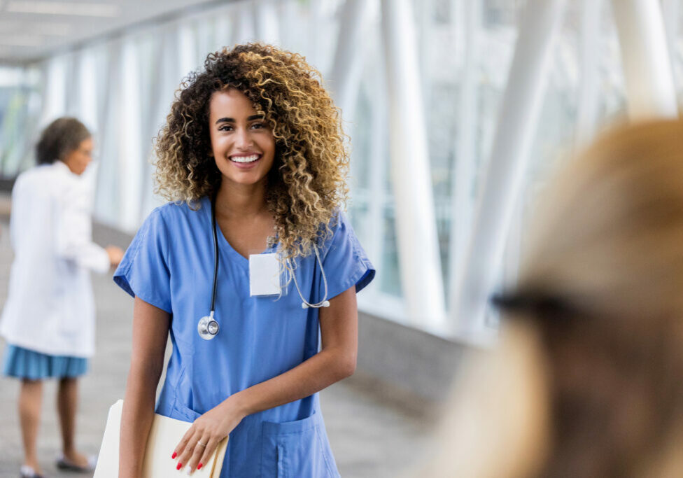 Beautiful mixed race young adult female nurse smiles at the camera while standing in a hospital hallway.