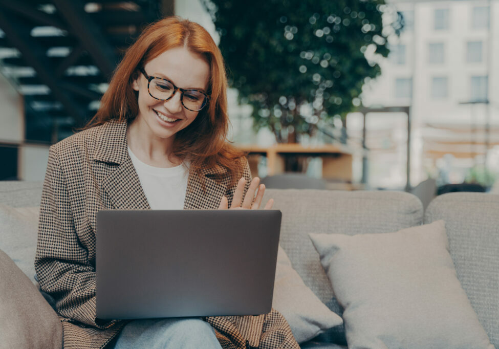 Happy red-haired business woman in optical eyewear waving at laptop webcam while sitting on sofa and having web conference with client, engaged in video call with colleagues while working remotely