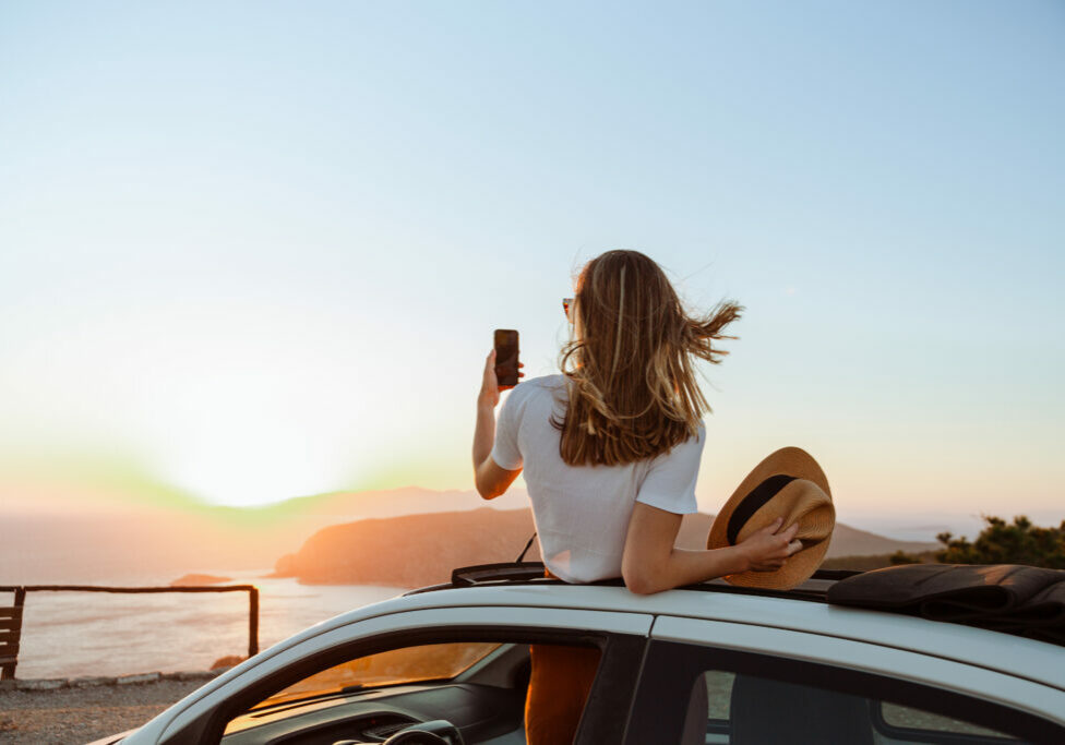 A photo of an unrecognizable woman looking at the sea and mountains from the opened roof of her car. She is taking photos of a beautiful sunset.