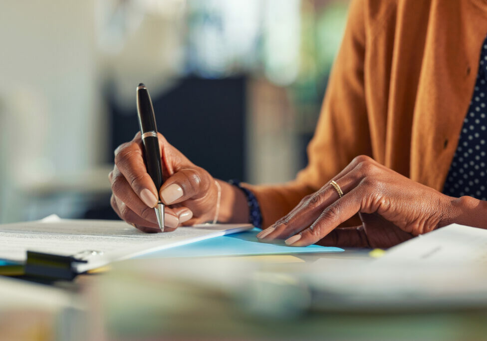 Close up of african american business woman hands writing on report while working in office. Successful black woman signing documents at home using a pen. Mid adult freelancer signing contract document on table.