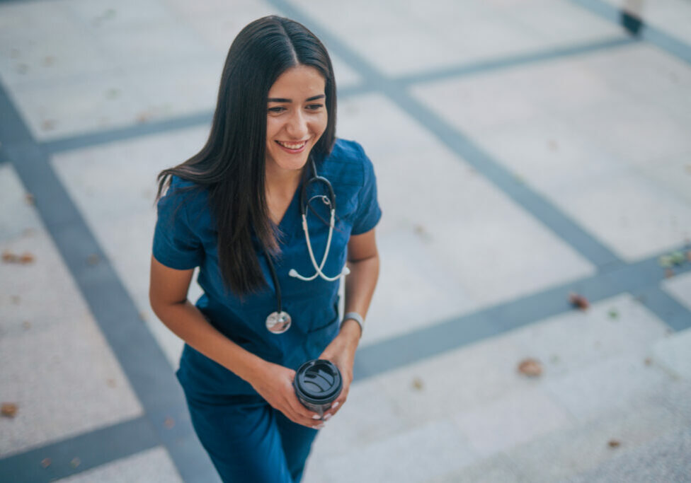 A young female nurse is walking outside the hospital, holding a cup of coffee in her hand